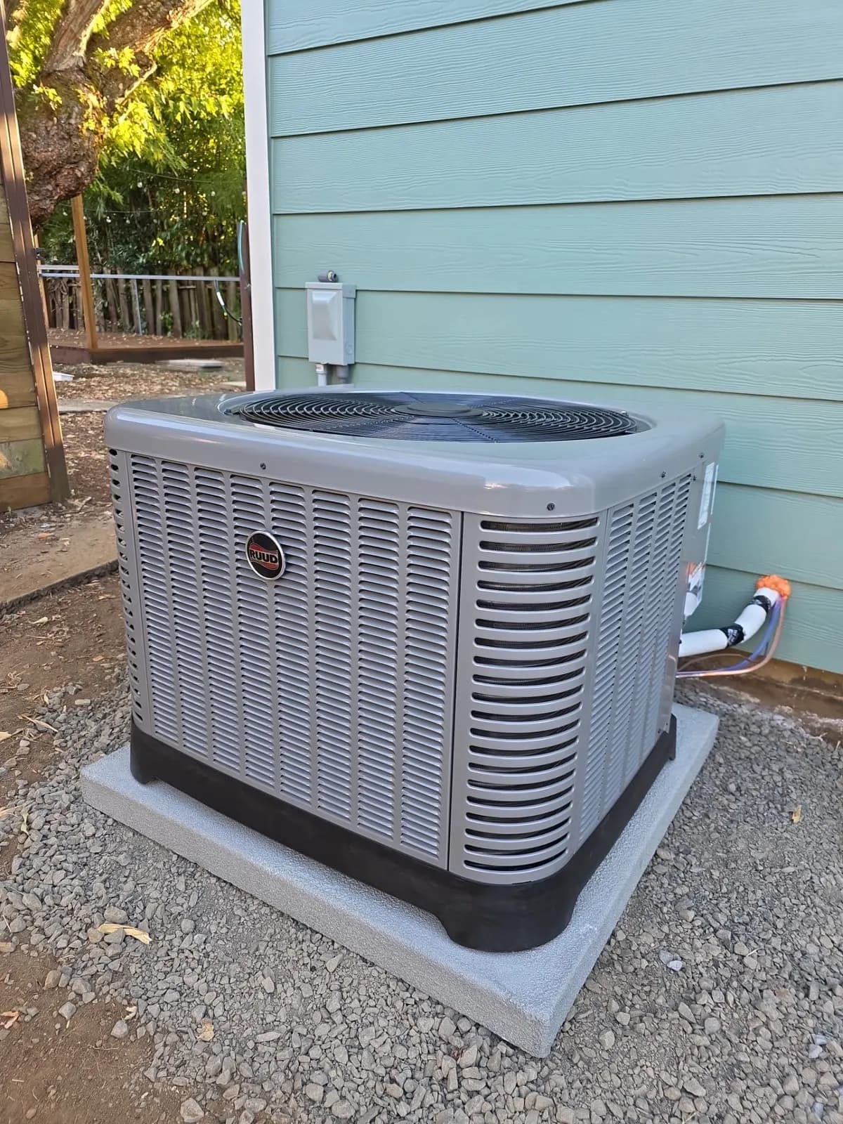 New gray air conditioner condenser on a concrete pad over gravel beside teal siding