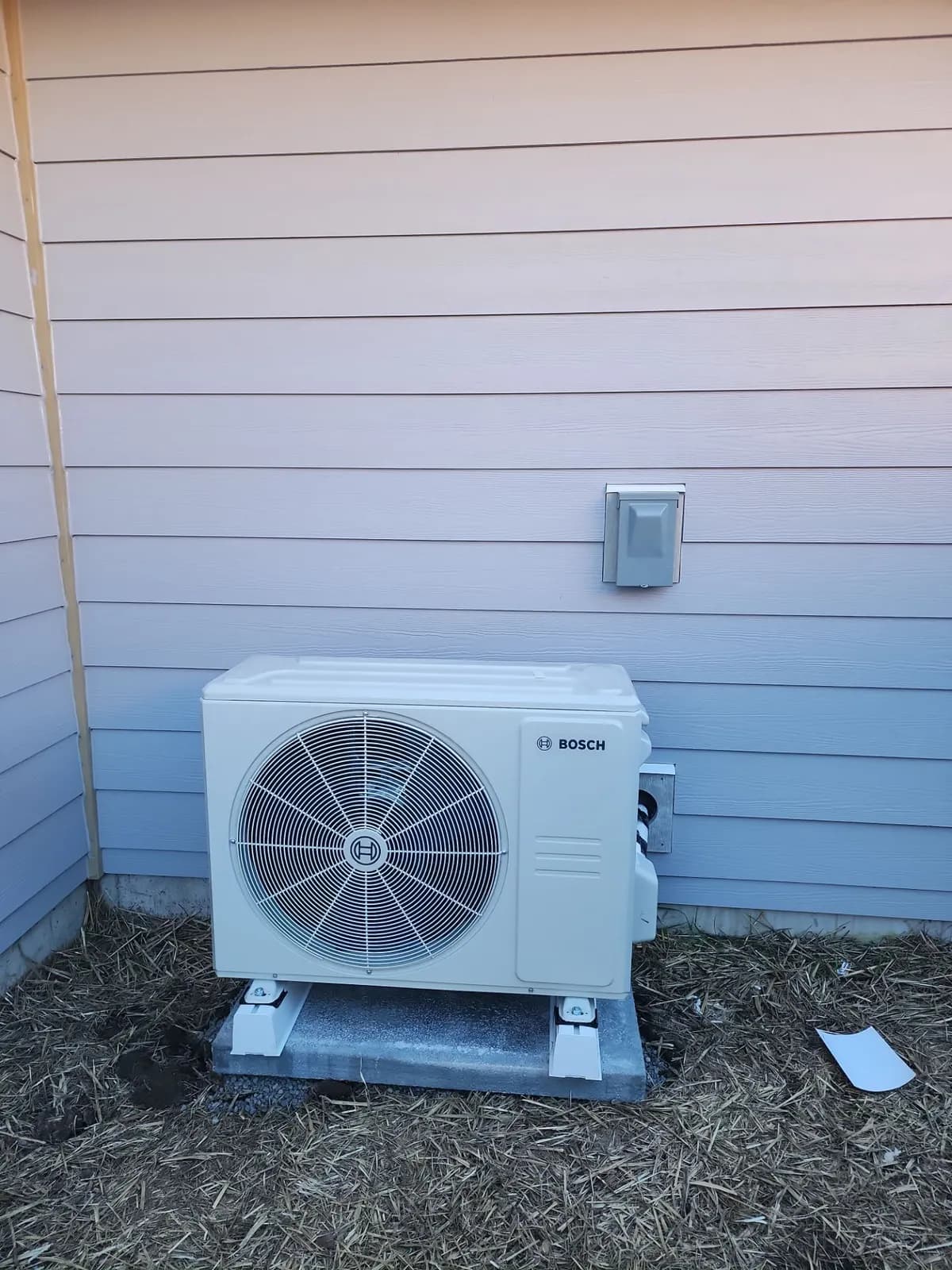 Bosch ductless mini split outdoor unit on mounting feet on a concrete pad beside light siding