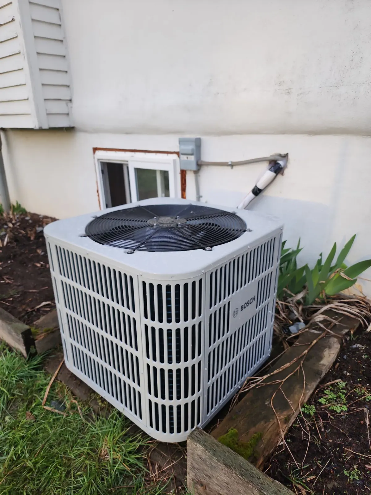 New white Bosch outdoor condenser beside a house with a wall-mounted disconnect