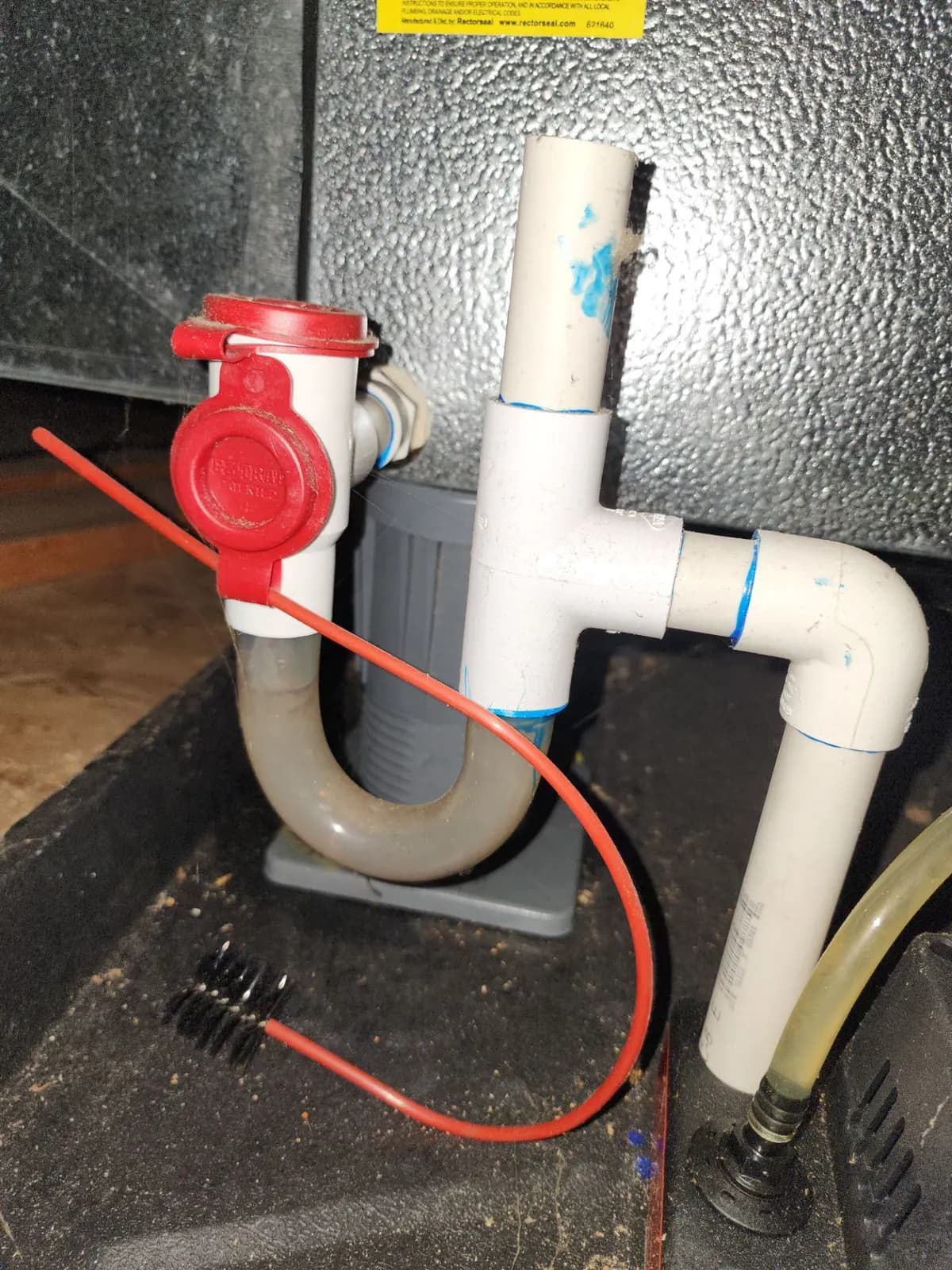 PVC condensate drain trap with a red cleanout cap and a cleaning brush under an indoor unit