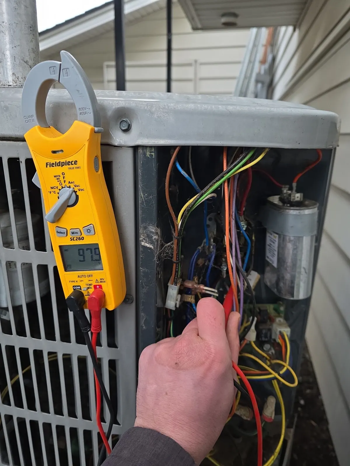 Technician testing wiring in an open condenser control panel with a clamp meter hung on the unit