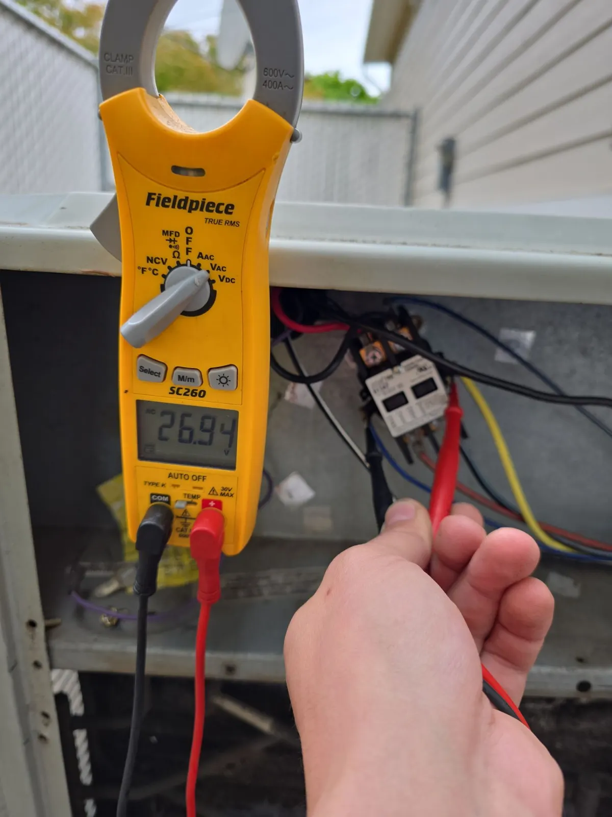 Technician probing the contactor in an open AC condenser with a clamp meter reading voltage
