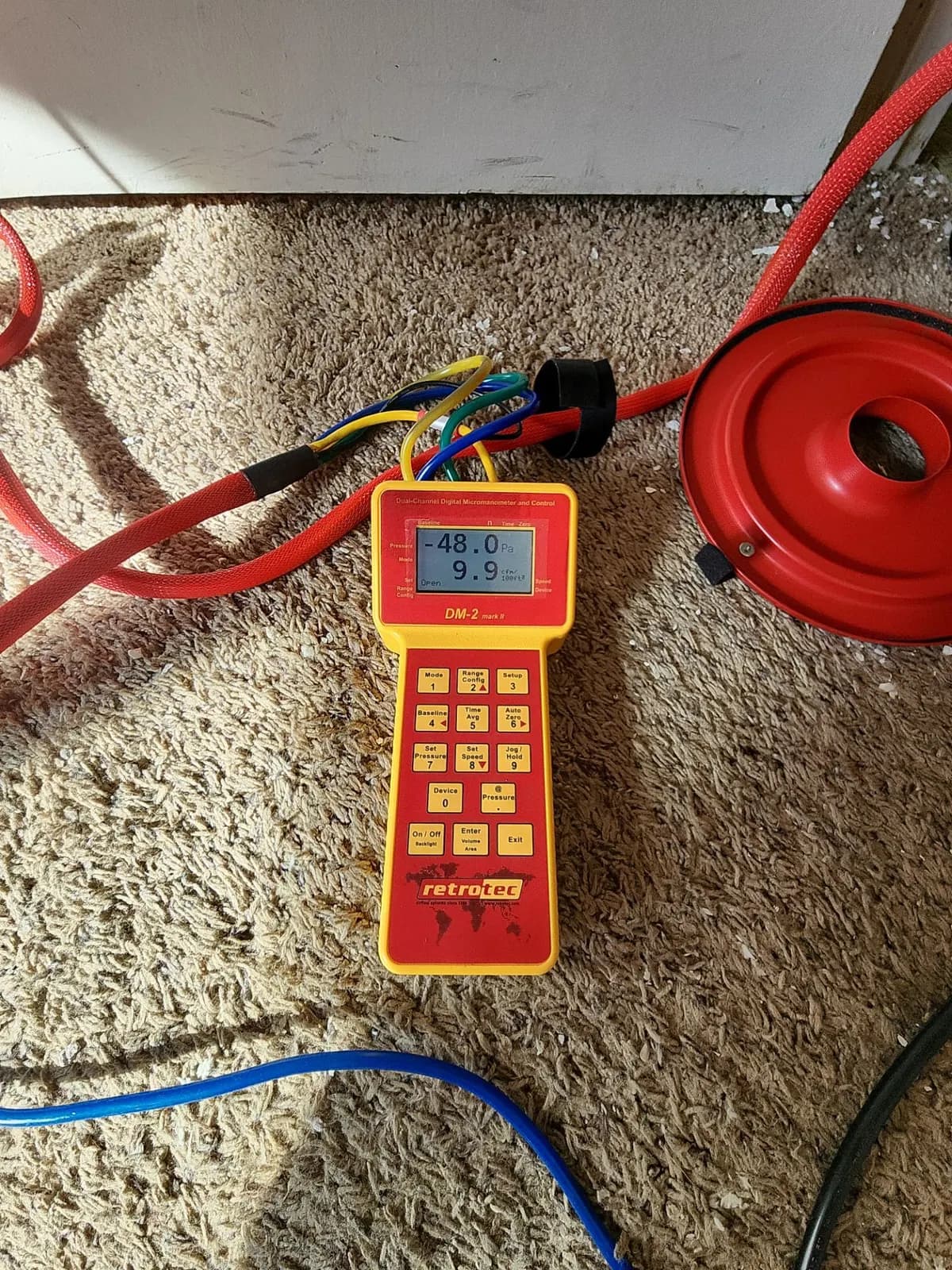 Digital manometer connected to a red duct test fan and hoses on carpet