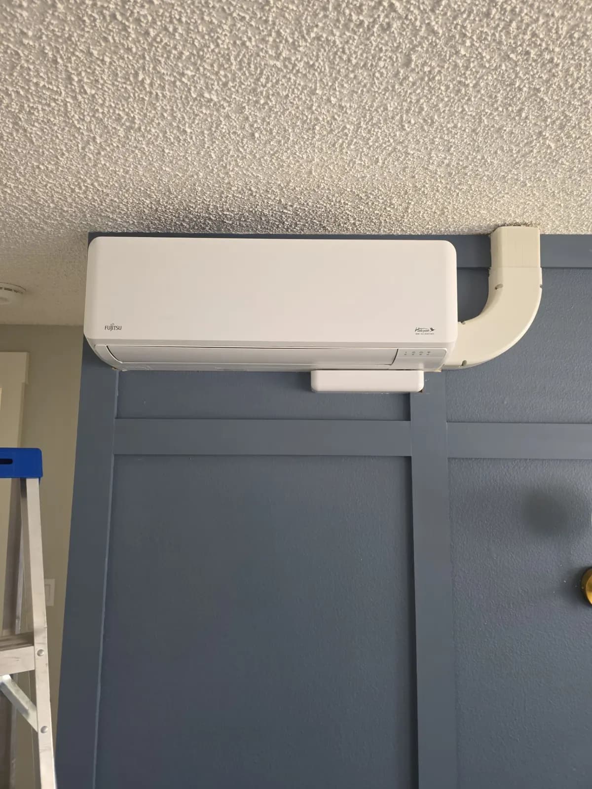 White ductless mini split head mounted near the ceiling on a dark paneled wall with a line set cover