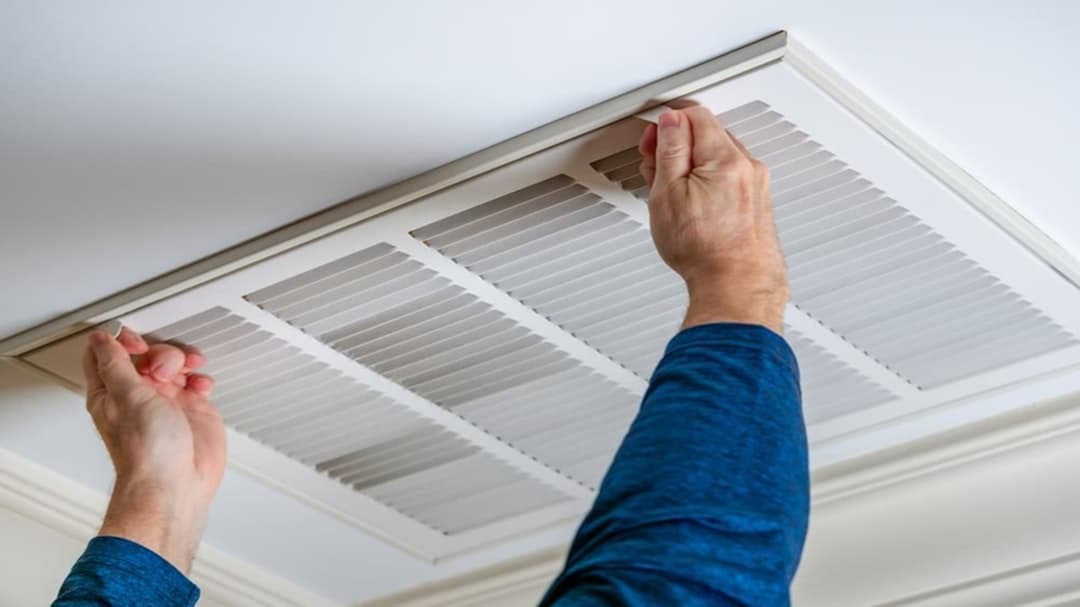 A person removing a ceiling return air grille to reach the filter behind it