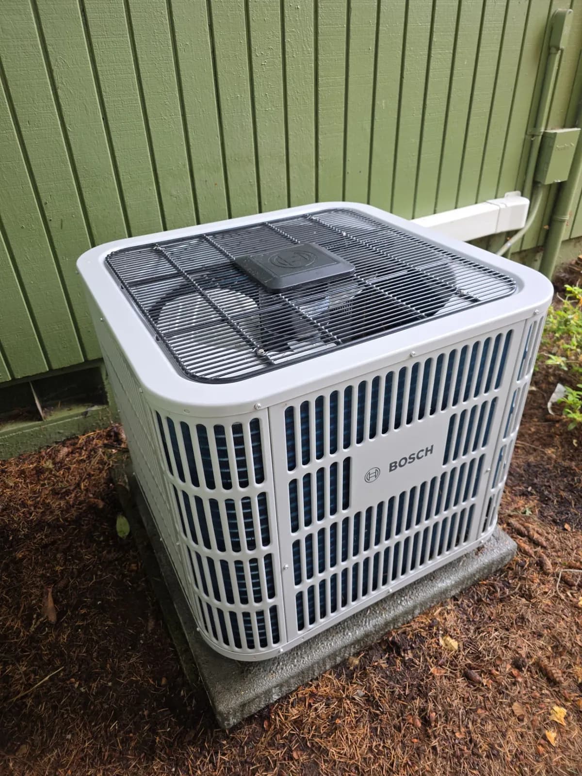New white Bosch heat pump outdoor unit on a pad beside green siding